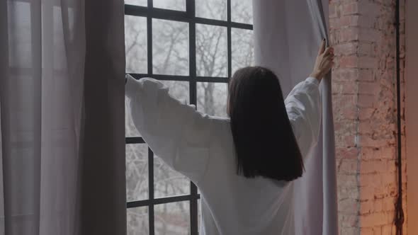 Young Woman Holding Curtains and Looking Through Window