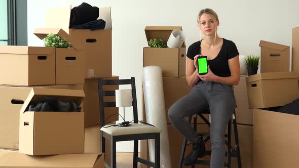 A Moving Woman Sits on a Chair in an Empty Apartment, and Shows a Smartphone with a Green Screen