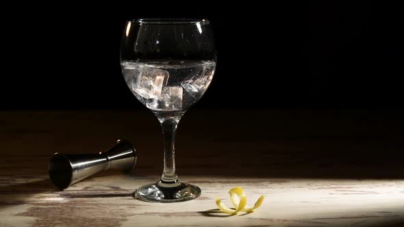 Person pouring ice cubes into glass of gin tonic with lemon and jigger on table in dark room