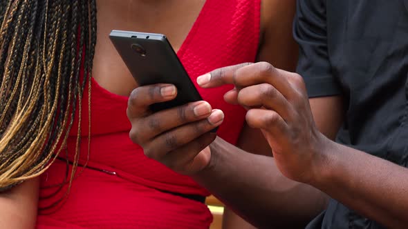 A Black Woman Teaches a Black Man Something on a Smartphone As They Sit on a Bench - Closeup