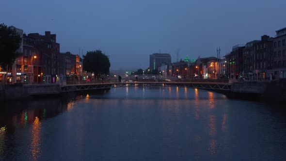 Pedestrians Walking on Footbridge Across River in Evening City