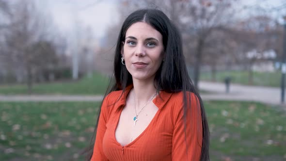 Young woman looking at the camera and smile while standing outdoors in the park in a windy day.