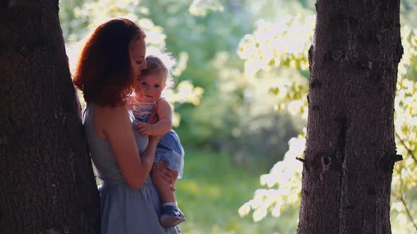 baby and mother are playing standing in the forest by a tree trunk in backlight