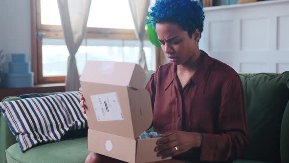 Young African American Woman Opens Cardboard Box and Upset Sits on Sofa