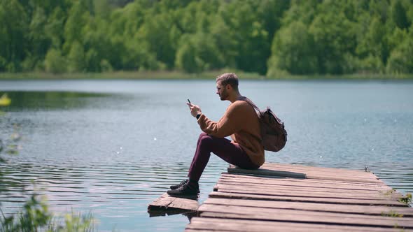 A Young Bearded Man with Glasses and a Backpack Sits on a Wooden Lake Pier in Slow Motion and Prints