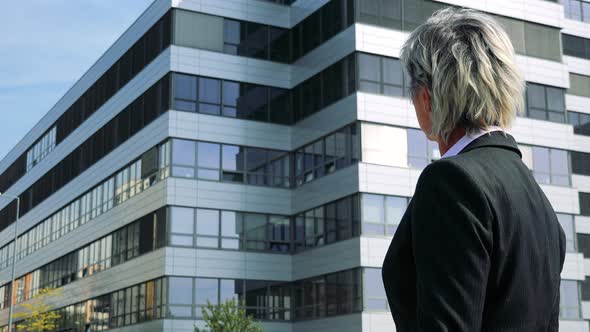 Business Middle Age Woman Looks Around (Shot From Side) - Company Building in the Background