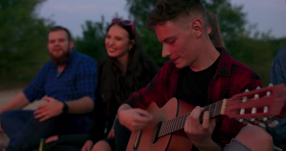 Cinematic View Through Bright Golden Flame on Young Teenage Man Playing Guitar Near Bonfire Smiling