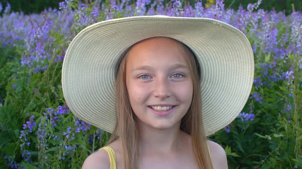 Smiling Girl in Hat Looking To Camera on Violet Lupin Meadow Landscape. Happy Girl Teen Portrait