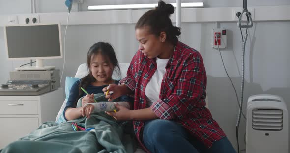 African Adoptive Mother and Asian Daughter Painting Hand Gypsum Together in Hospital Ward