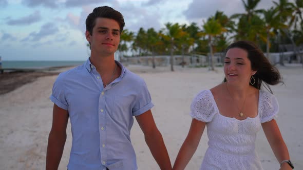 Romantic young adults walk on beach. Tropical palm trees and white sandy beach. Evening sunset and w