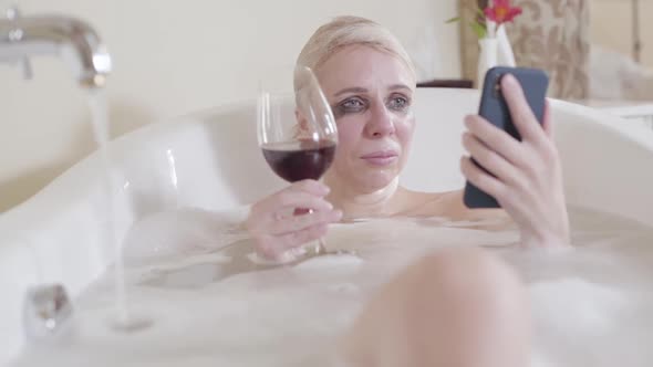 Crying Blond Woman Reading Messages on Smartphone and Drinking Red Wine. Portrait of Depressed