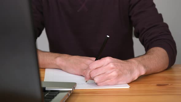 Left-handed Man Writing On A Paper In Front Of A Laptop On The Table. close up