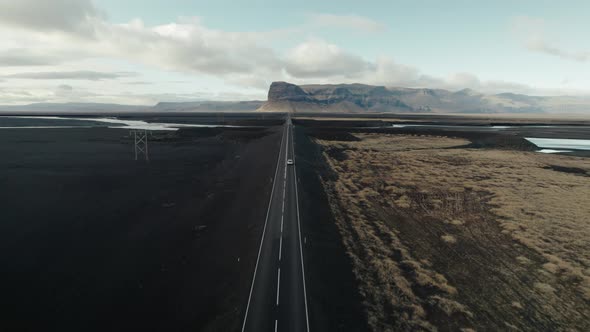 Car drives long straightaway through black sand desert in Iceland. Aerial follow shot.