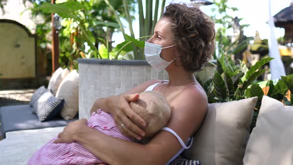Young Caucasian Woman in a Face Mask Breastfeeds Her Baby Daughter in a Garden Restaurant