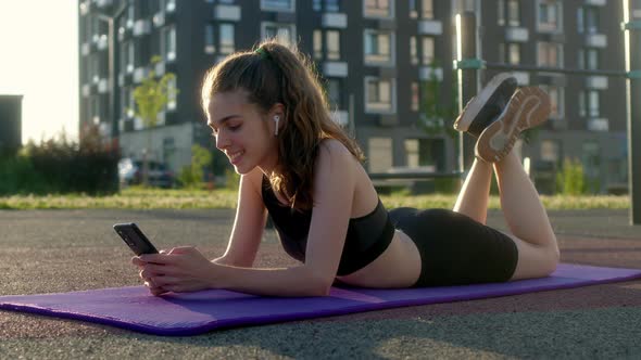 Cheerful Happy Young Woman Lies on Fitness Mat During Break Between Outdoor Sports and Communicates