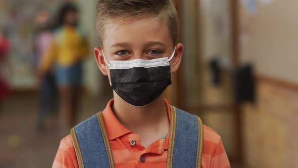 Portrait of caucasian schoolboy wearing face mask, standing in corridor looking at camera