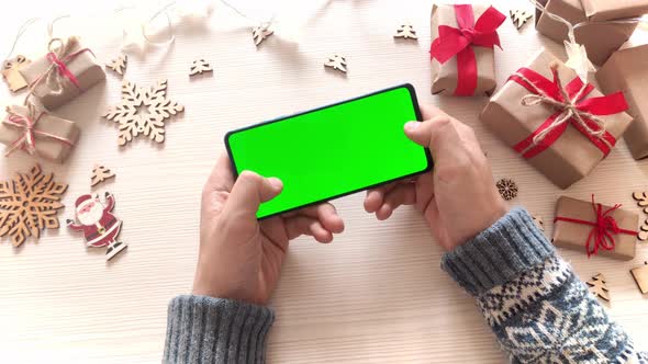 Man holding smartphone with horizontal green screen and touch on Christmas background with gifts.