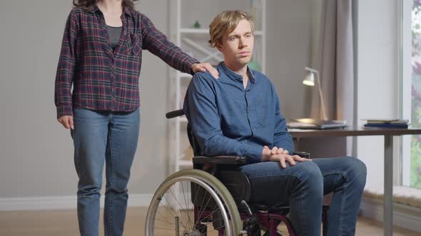 Sad Depressed Disabled Man in Wheelchair Thinking As Unrecognizable Woman Putting Hand on Shoulder
