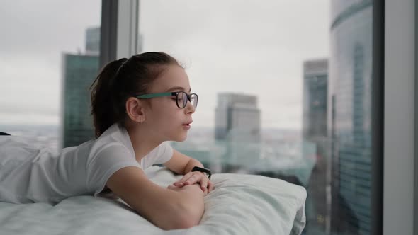 Cute Teenage Girl Relaxing on a Comfortable Bed with a White Blanket Enjoying the City View From