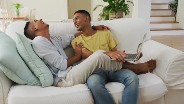 Smiling mixed race gay male couple sitting on sofa using laptop and talking