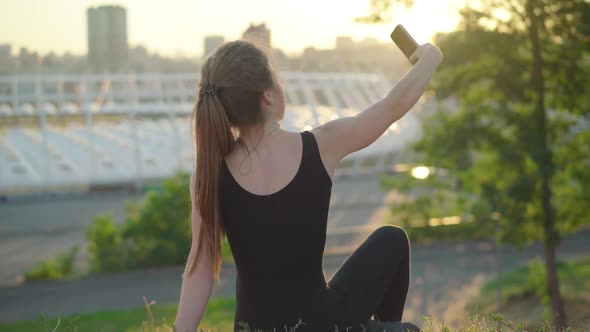 Back View of Young Caucasian Woman Sitting on Park Hill at Sunset and Taking Selfie. Carefree Slim