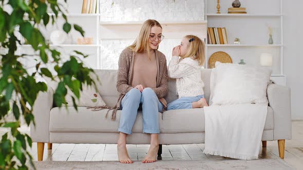 Adult Mother Sits on Sofa at Home in Living Room with Little Girl Daughter Covering Mouth with Hands