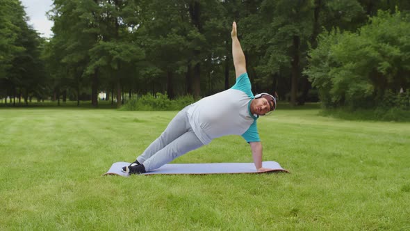 Portrait of Stout Man in Headphones Doing Yoga Exercises on Fitness Mat Outdoors