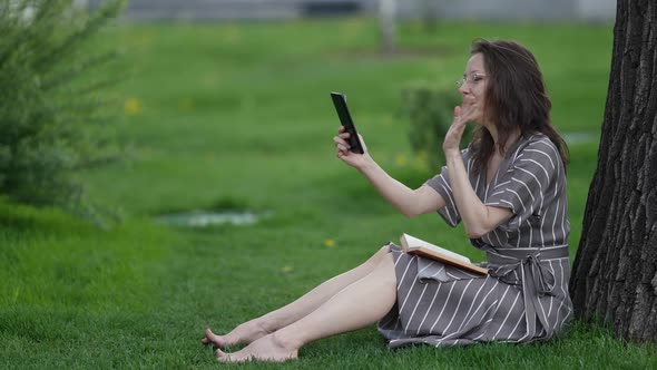 Adult Townswoman is Resting in Park Sitting on Grass and Talking By Cellphone