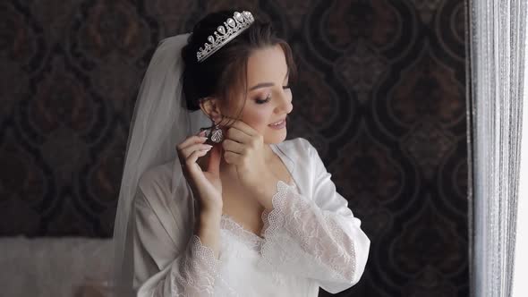 Bride in Boudoir Dress Puts on Earrings. Wedding Morning Preparations. Woman in Night Gown and Veil