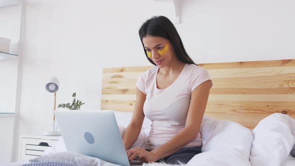 Сheerful Young Woman Working at Home Using Laptop in Bed in the Morning with Collagen Eye Patches