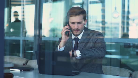 Young Businessman in a Suit Is Sitting on the Terrace of a Cafe and Working on a Laptop, Talking on