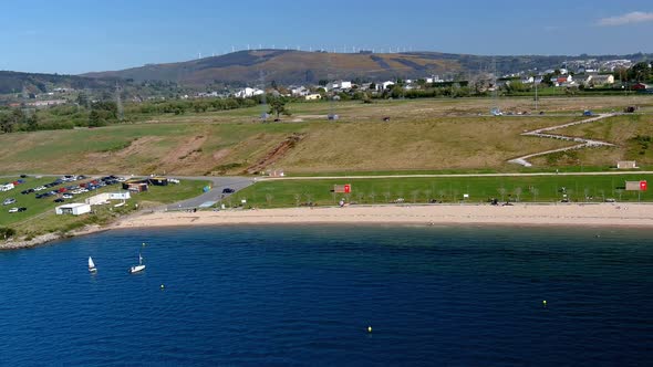 people on the beach and sailing on the lake with the village behind and the wind turbines at the bot