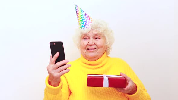 Older Woman with a Gift Wear Yellow Sweater and Horn Cap on a White Background Holding Smartphone