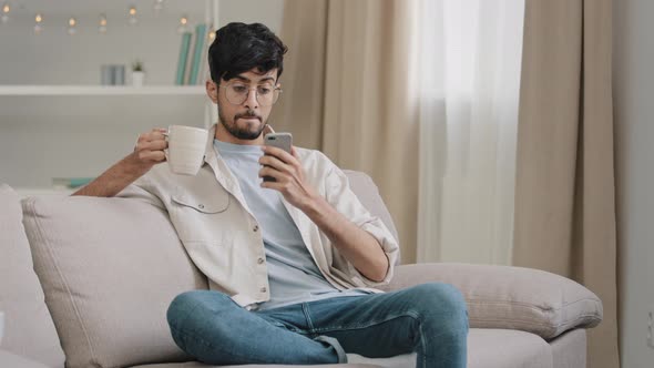 Millennial Arabic Guy Indian Handsome 30s Man Sitting at Home on Couch Drinking Hot Tea Coffee