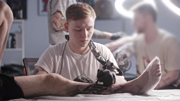 A Session at the Tattoo Salon Young Man Master Tattooing Big Letters on the Leg Looking in the