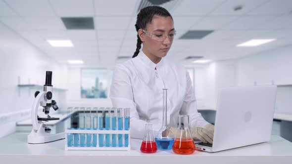 A Female Scientist Writes the Results of a Genetic Study To a Laptop While Sitting at a White Table