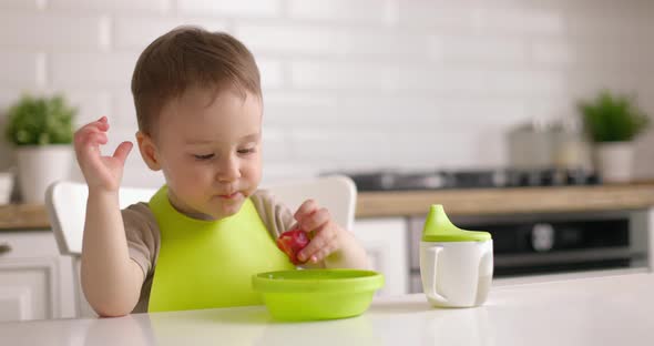 Cute Little Baby Boy Sits at a Table in the Kitchen and Eats Strawberries From Green Plate with His