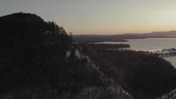 Curving around the ridge of a forested mountain at dawn to reveal a steep cliff AERIAL