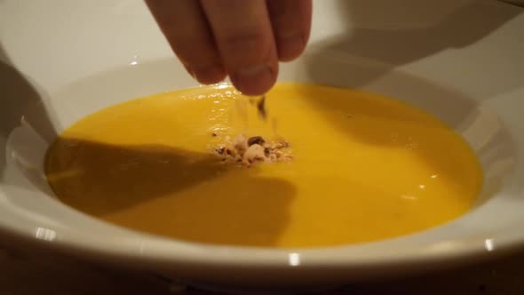 A white male hand, model is sprinkling nuts on a yellow soup in a white bowl.