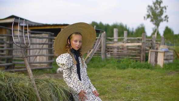 Rustic Girl in Vintage Dress and Hat on Haystack and Pitchfork Background in Countryside