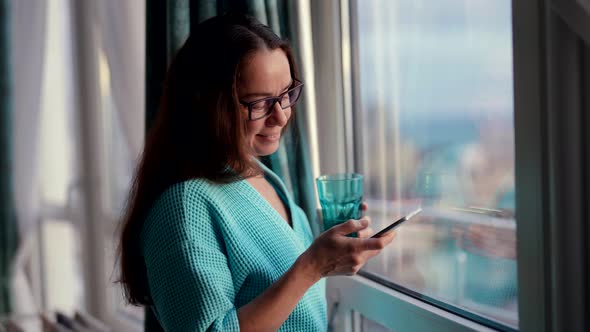 Pretty Middleaged Woman is Sending Message By Apple in Smartphone and Drinking Juice Near Window