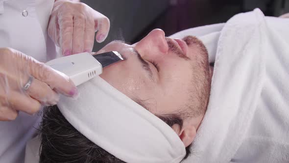 Ultrasonic Face Scrubber. Beautician Cleans the Face of a Young Man