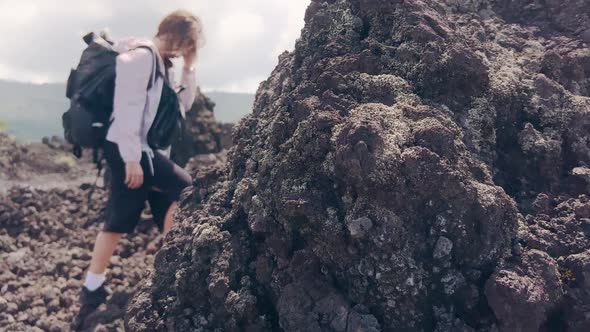 A Traveler Girl with Sunglasses and Two Backpacks Approaches a Stone Block of Black Lava on Which a