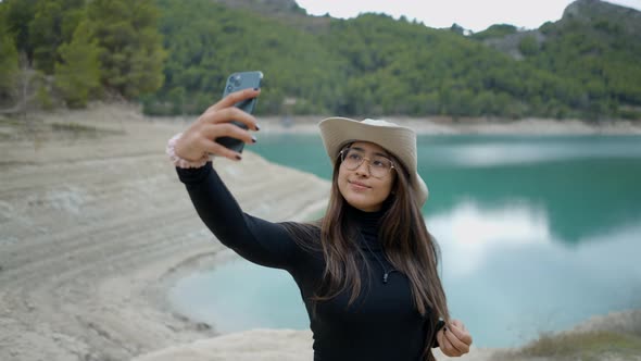 Darkhaired Girl in Hat and Glasses Takes Selfie By Lake Close View