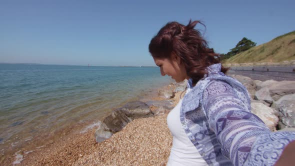 POV Shot Of Young Woman Reaching Down To Pick Up Plastic Bottle On Beach