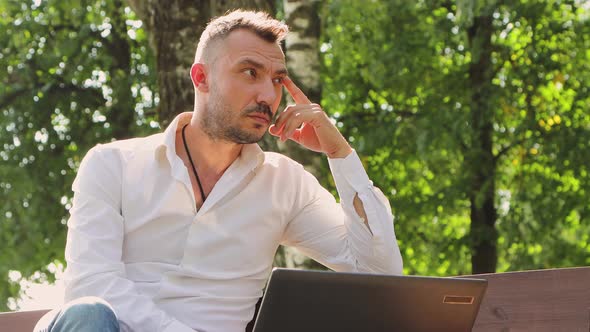 A Young Man in a White Shirt is Thoughtfully Talking Online on a Laptop