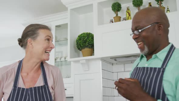 Smiling senior diverse couple wearing blue aprons and cooking in kitchen