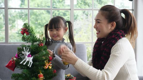 Asian mother and daughter in sweather, decorating christmas tree together in living room