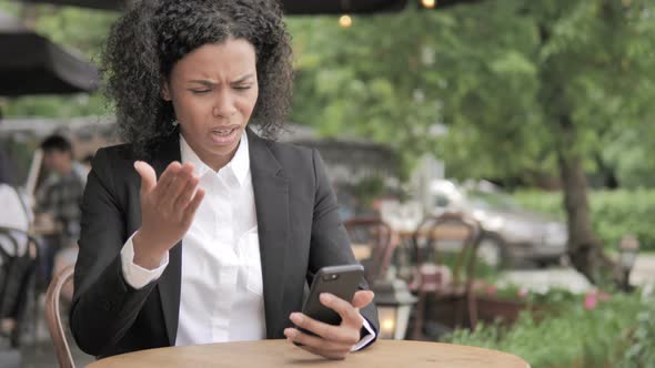 African Woman Upset by Loss on Smartphone, Sitting in Outdoor Cafe