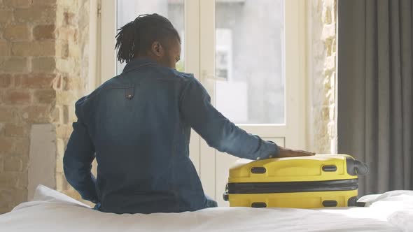 Back View of African American Young Man Sitting on Bed with Luggage Before Trip, Taking Yellow